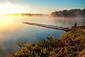 Man on a wooden boardwalk pier photographing a tranquil, fog-covered lake at sunrise