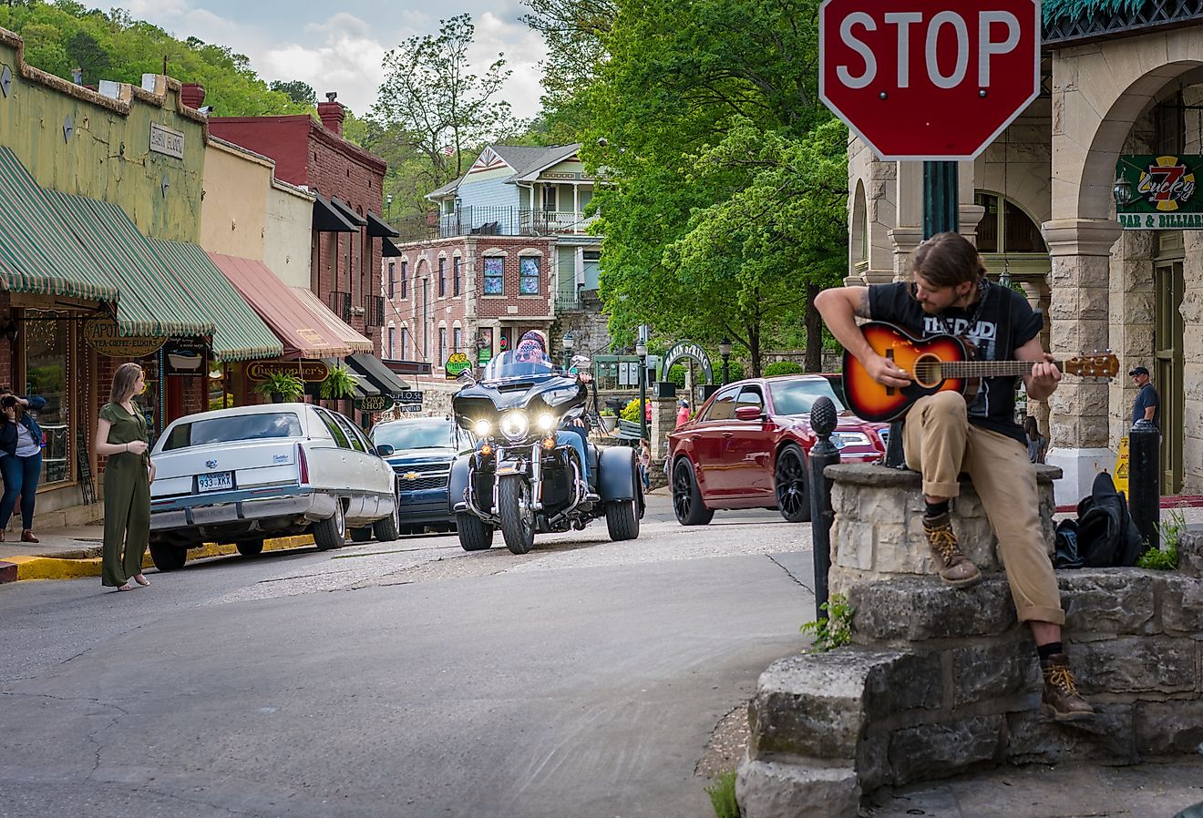 Man playing guitar at a stop sign, and cars and people on the street in downtown Eureka Springs, Arkansas. Image credit shuttersv via Shutterstock