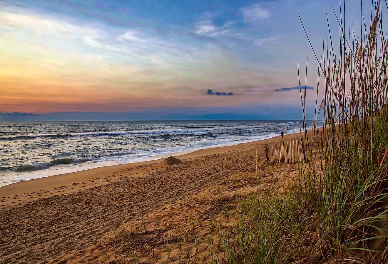 Colorful sunrise on a North Carolina beach, waves breaking on a sandy shore framed by dune grass. Duck, NC. Image credit Jeremy Tyree via Shutterstock. 