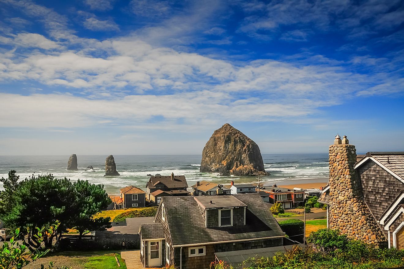 The town of Cannon Beach, Oregon, with the Haystack Rock. Coastal village with houses overlooks the ocean where a large, rugged rock formation rises from the water. The sky is blue with scattered clouds.