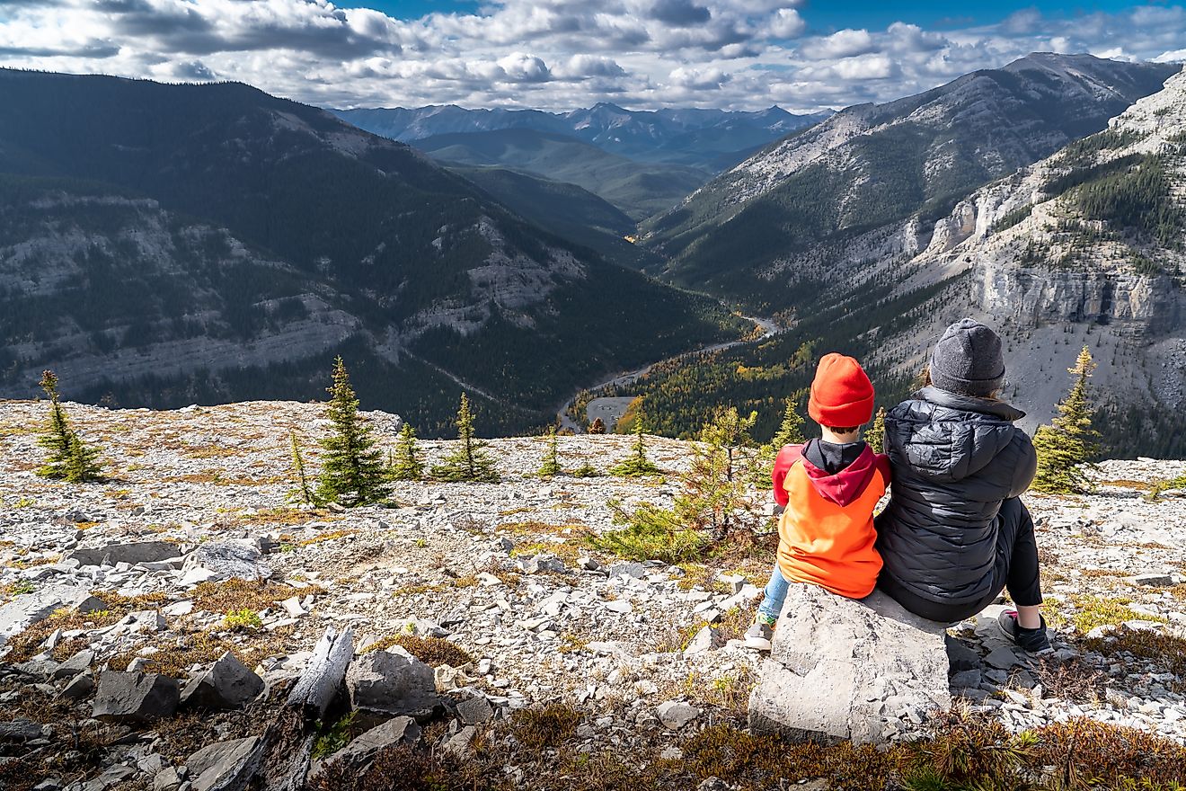 A brother and sister sitting on a rock overlooking the Canadian Rocky Mountains, with a valley of trees displaying fall colors on Moose Mountain near Bragg Creek, Alberta, Canada.