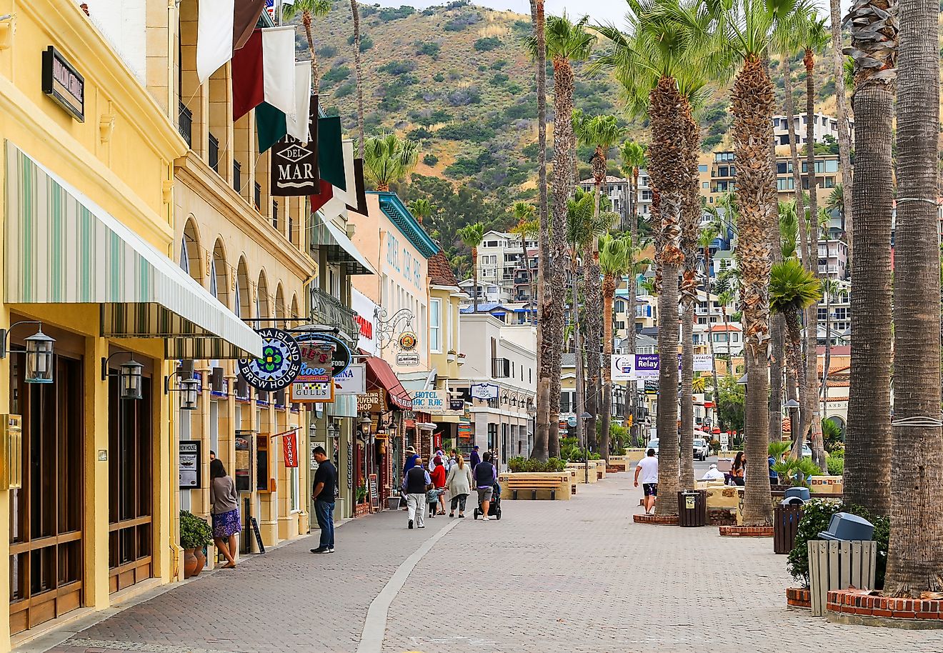 The boardwalk in Avalon (Santa Catalina Island) with shops on the left. People strolling around. In the background are houses in the hills.