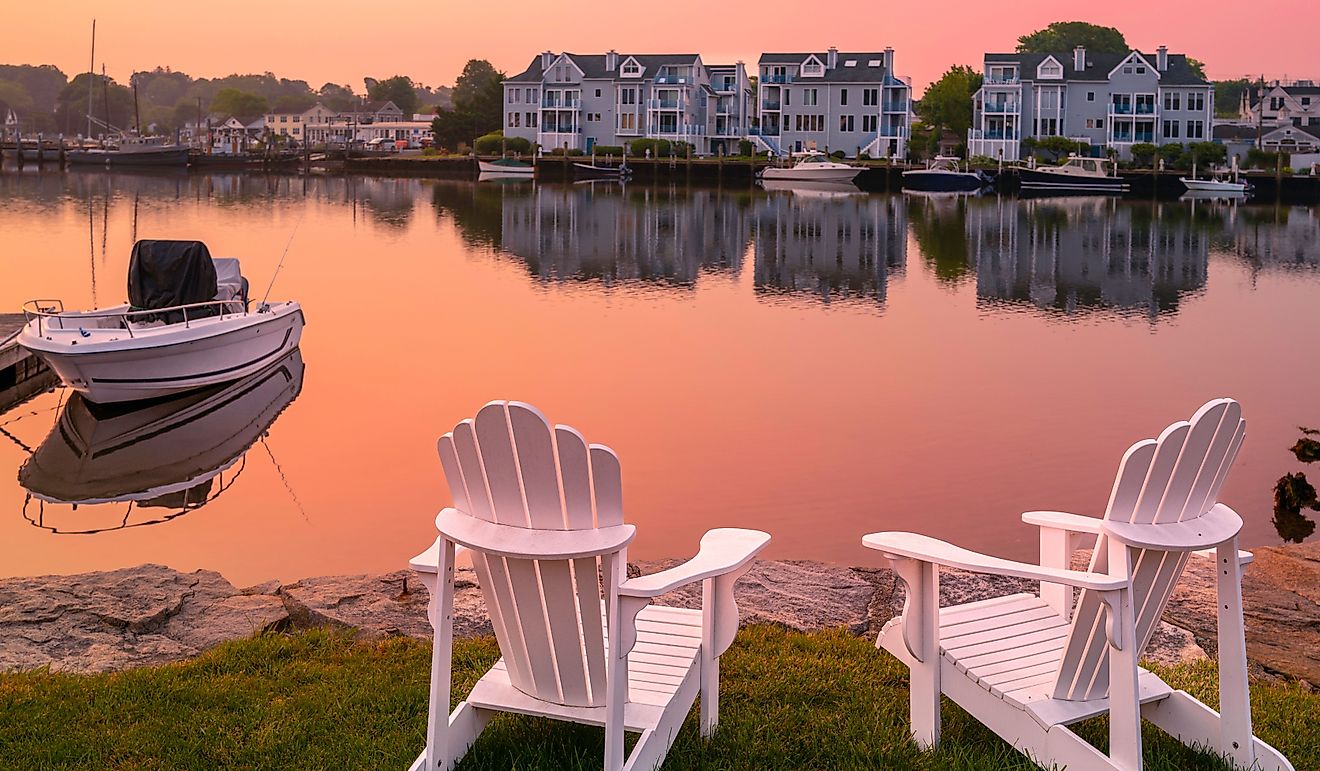 Sunrise, two white Adirondack chairs on the beach, a moored boat, and wildfire smoke over the Mystic River marina village in Connecticut.