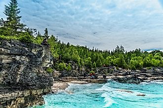 Backpackers taking a break on limestone cliffs before the rolling waves of Georgian Bay. A dense coniferous forest and overhead clouds form the backdrop. Image by Golden Shrimp via Shutterstock.com