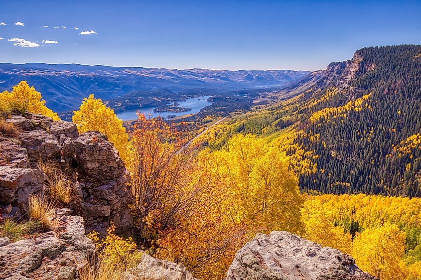 View into a valley with bright yellow aspen trees and a lake with rock walls in the foreground on a sunny day near Durango, Colorado.