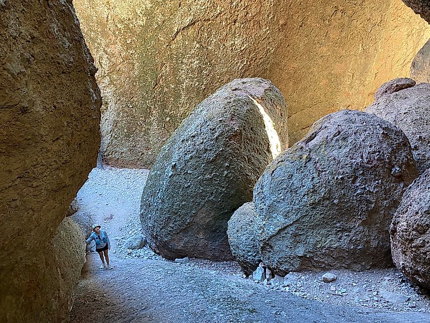 A female hiker stands at the bottom of a steep hill leading out of a large cavern with massive boulders.