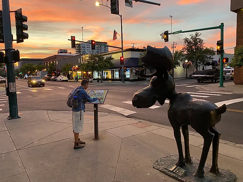 A woman stops to read an interpretive sign next to a large bronze moose statue. A stunning, post-sunset sky is seen overtop of the commercial street.