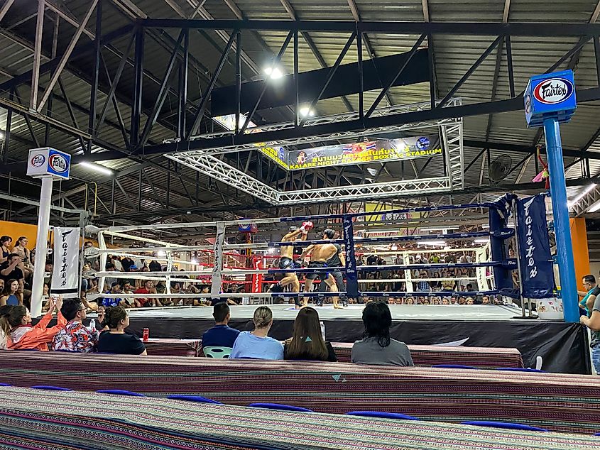 A muay thai fight in a small stadium. One of the fighters is winding up for a kick against the other. 