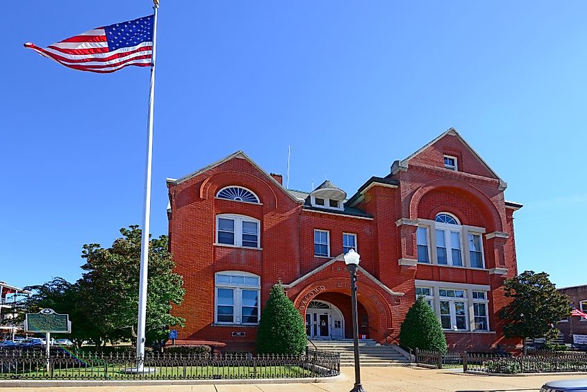 A historic red-brick courthouse building in Oxford, Mississippi, featuring arched windows and a central entrance. An American flag flies on a tall flagpole in front, with a small fenced lawn and trees under a clear blue sky.