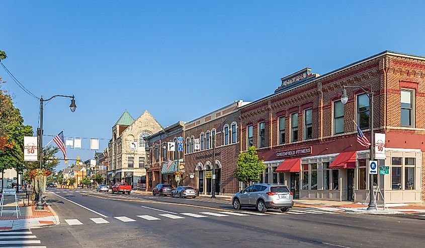 A quaint small-town street with historic brick buildings, American flags, parked cars, and clear blue skies. A calm, inviting atmosphere.