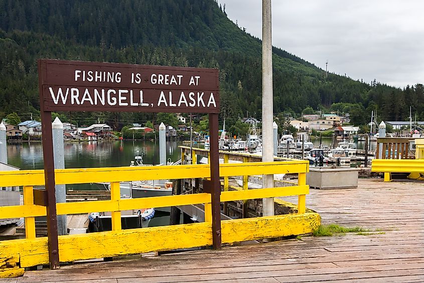 A sign reads "Fishing is Great at Wrangell, Alaska" on a dock with yellow railing. Boats are moored in a scenic harbor, surrounded by lush green hills.