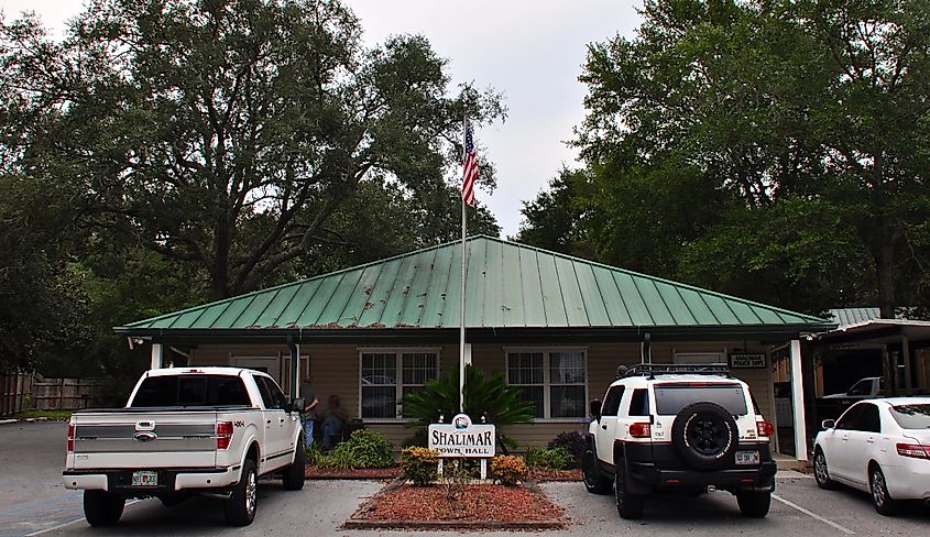 Shalimar Town Hall in Florida on a cloudy day, with a jeep, a pickup truck, a sedan, and two people out front.