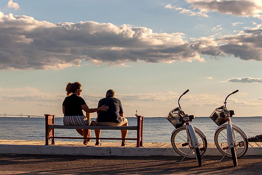 A middle-aged Caucasian couple sits on a bench by the beach in Rock Hall, MD, with their identical bikes parked nearby. The woman is giving the man a pat on the back.