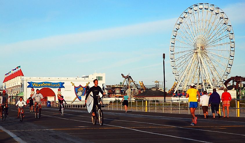 An adult man rides his bicycle while carrying his surfboard in the boardwalk in front of a Ferris Wheel, in Ocean City, New Jersey.