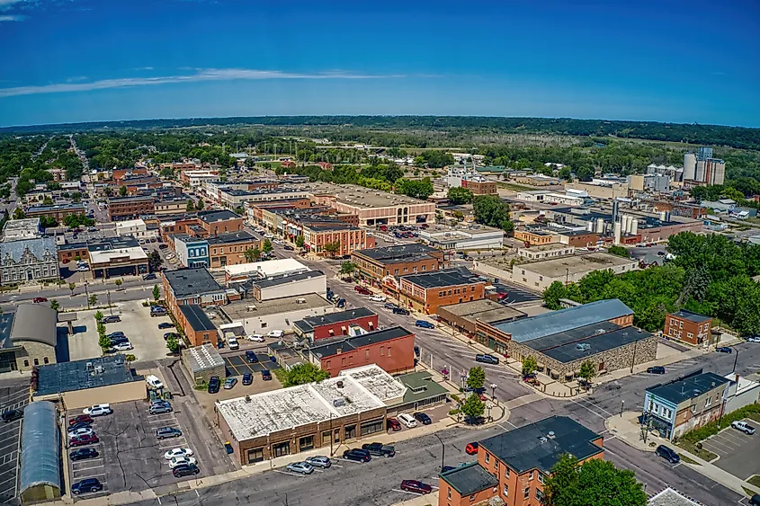 Aerial view of a small town with brick buildings, tree-lined streets, and parking lots. The horizon shows lush greenery under a clear blue sky.