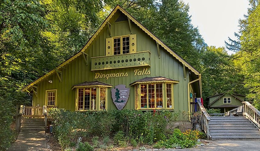 Green cottage-style visitor center labeled "Dingmans Falls," surrounded by lush trees and colorful flowers, evoking a peaceful, inviting atmosphere.