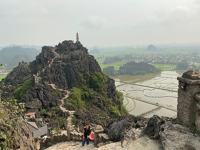 A woman pauses on a long slight of stone stairs, looking up toward the top of a mountain while a sister mountain with another staircase stands tall in the background.