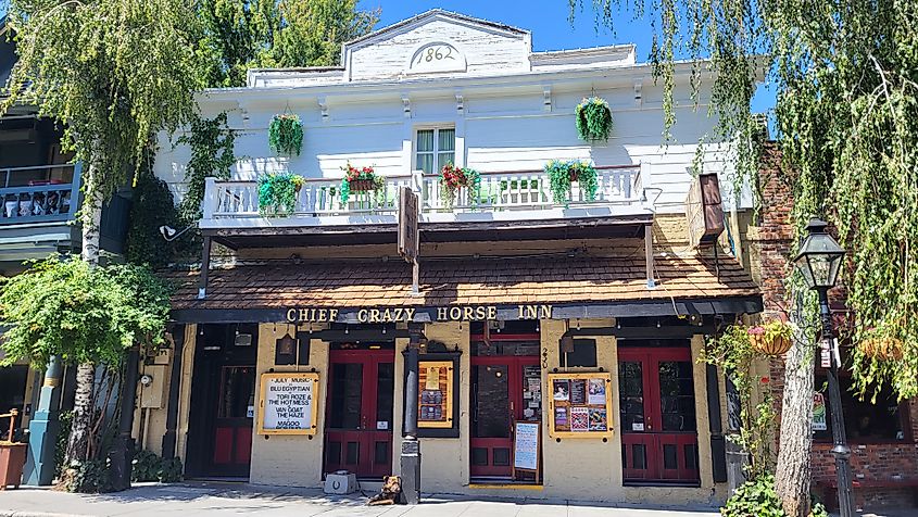 Historic two-story inn with wooden façade, named "Chief Crazy Horse Inn," features red doors, lush greenery, and vibrant hanging plants under a clear blue sky.