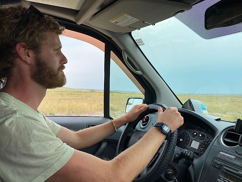 A blond, bearded man drives through a prairie setting. 