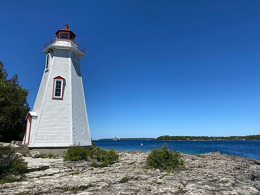 A large white lighthouse with red trim standing on a rocky shore underneath a bluebird sky.