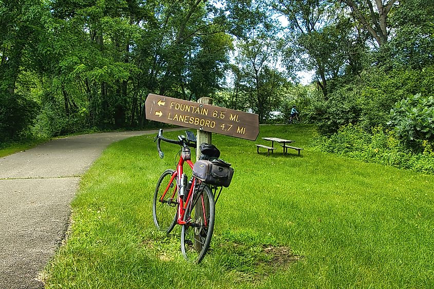 Red bicycle leaning against a wooden trail sign with directions to Fountain and Lanesboro. The path is surrounded by lush green trees and grass.