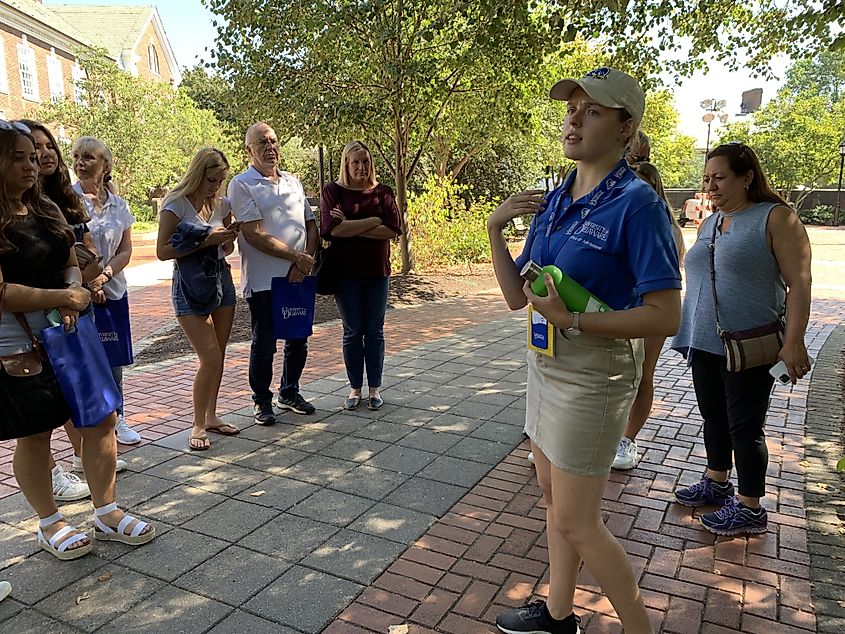 A student guide wearing a University of Delaware shirt speaks to a group during a campus tour, with visitors standing on a shaded walkway nearby.
