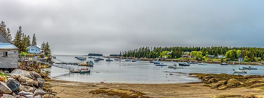 A fleet of Maine lobster boats moored in a calm bay on a gray, cloudy, and foggy day, with the boats barely visible through the mist, creating a quiet and serene atmosphere, in Stonington, Maine.