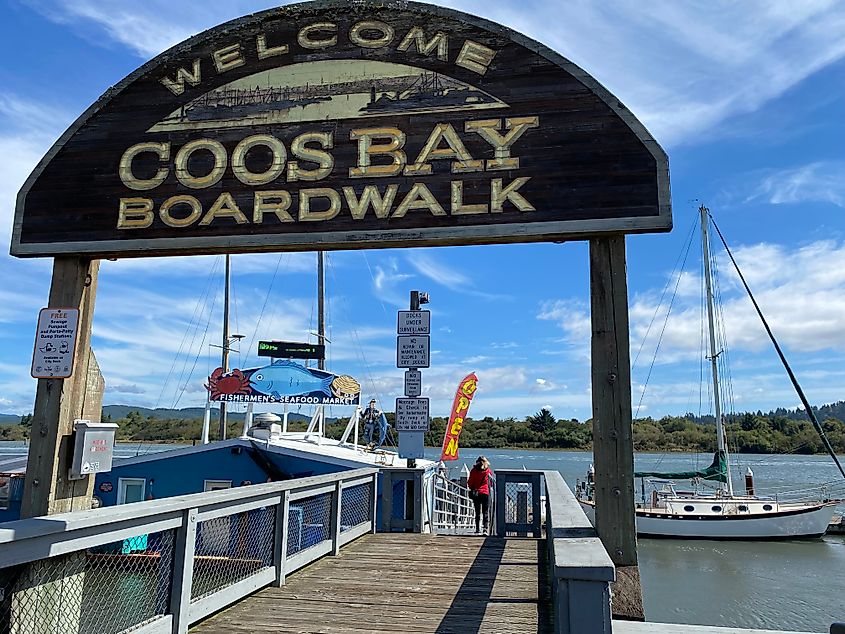 Wooden sign reads "Welcome Coos Bay Boardwalk" over dock. A person walks toward boats on a sunny day. Scenic waterfront with boats and trees.