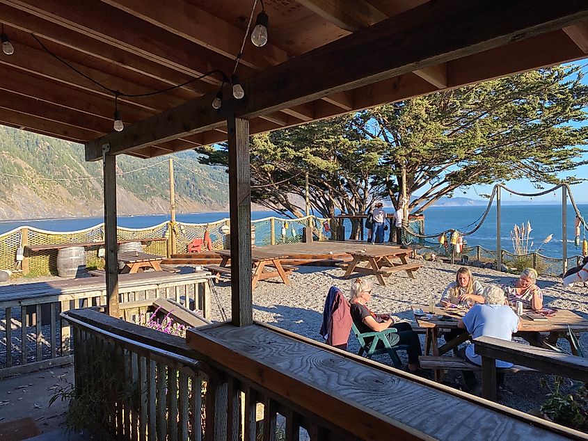 People gather around the picnic tables of an open-air seaside restaurant.  