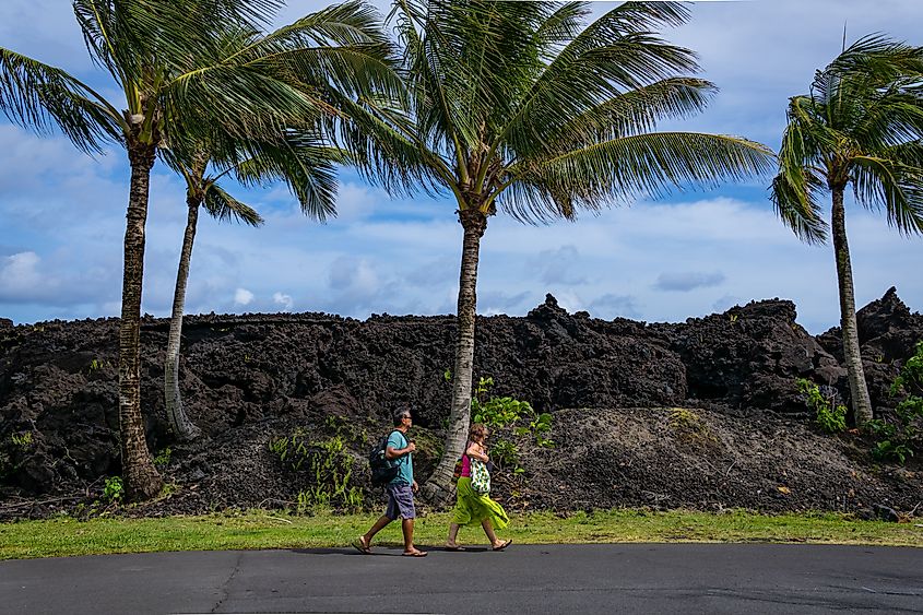 Editorial Photo Credit: Chris Allan via Shutterstock. PAHOA, HAWAII - APRIL, 29, 2023: Photo of two visitors walking past the black lava wall formed at Isaac Hale Pohoiki State Park formed by t 2018 eruption of the Kilauea volcano.