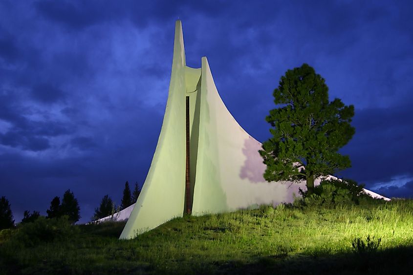 A modern, angular white structure with upward arcs sits on a grassy hill under a dramatic twilight sky. A lone green tree stands to its right.