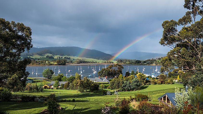 A double rainbow arches over the marina in Cygnet, Tasmania, with boats docked peacefully in the water, framed by the vibrant colors of the rainbow and the serene coastal landscape.