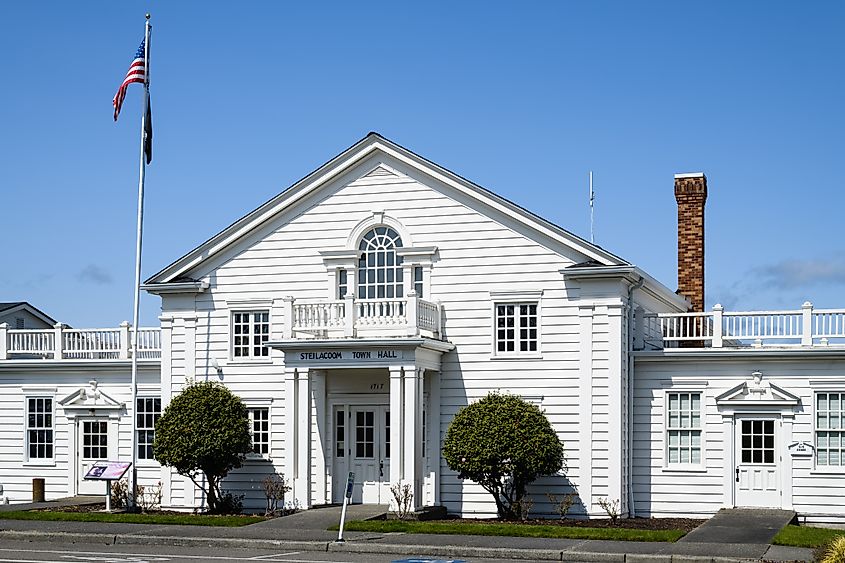 A white, colonial-style town hall with a gabled roof and large windows, flanked by green shrubs. A flagpole with the American flag stands in front.