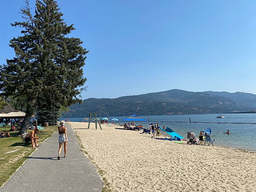 A young woman strolls along a sidewalk next to a sandy beach on a bluebird day.