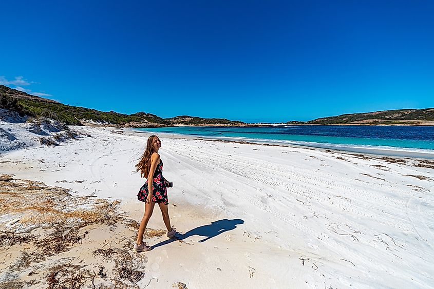 A long-haired girl in a flowing black dress adorned with roses walks along a pristine white sand beach with turquoise waters and striking orange rock formations at Cape Le Grand National Park near Esperance, Western Australia.