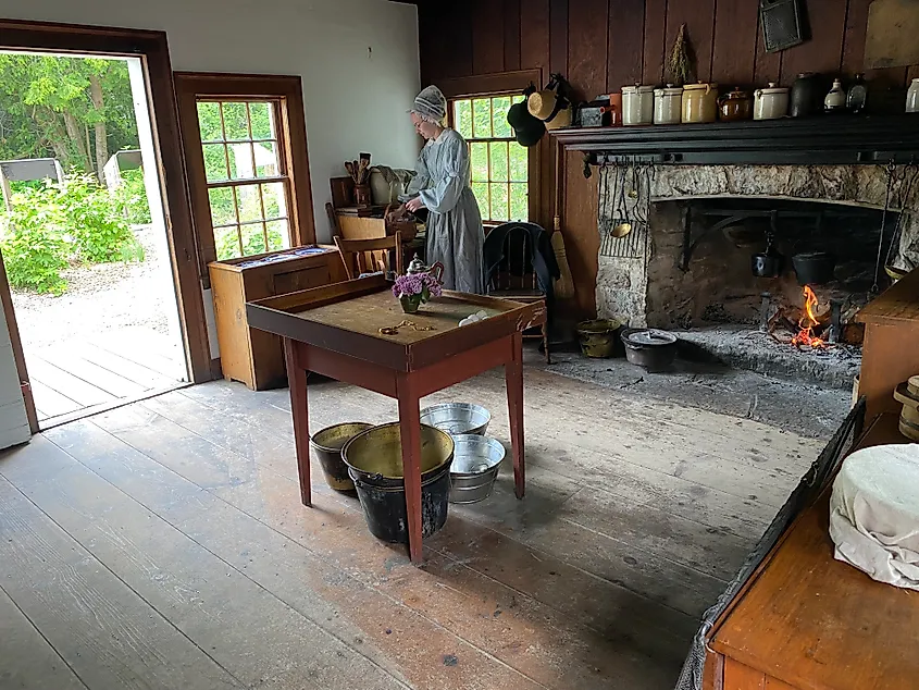 A period actress boils a cast iron pot over a fireplace in a quaint, 19th century home. 