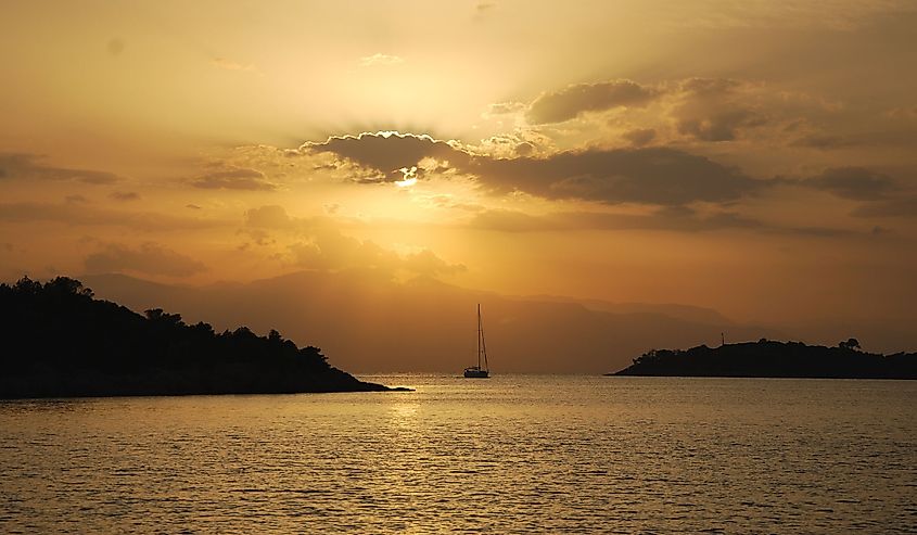 Sailing yacht entering Porto Kheli (Greece, Argolic Gulf) backed by a golden sunset and mountains in the distance.