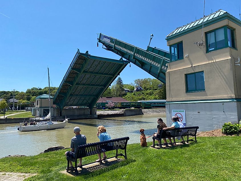 People on benches watching a green steel lift bridge open for a sailboat.