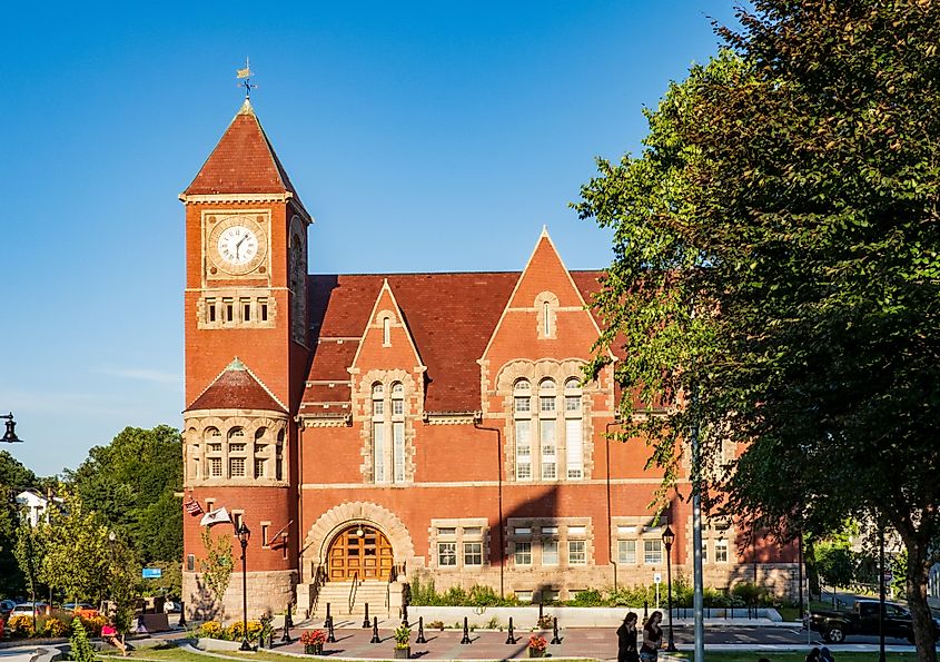 Red-brick historic building in Amherst, Massachusetts with a tall clock tower, arched entrance, and steep gabled roof, framed by green trees under a clear blue sky.