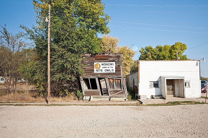 Dilapidated wooden building with a "Monowi Home of the Nite Owls" sign, next to a small white brick structure. Surrounding trees and clear sky in the background.