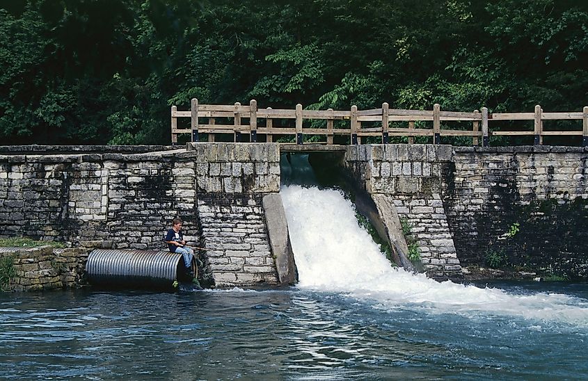 A stone bridge with water flowing rapidly beneath. A person sits on a metal pipe beside the stream, surrounded by lush greenery, conveying a serene atmosphere.