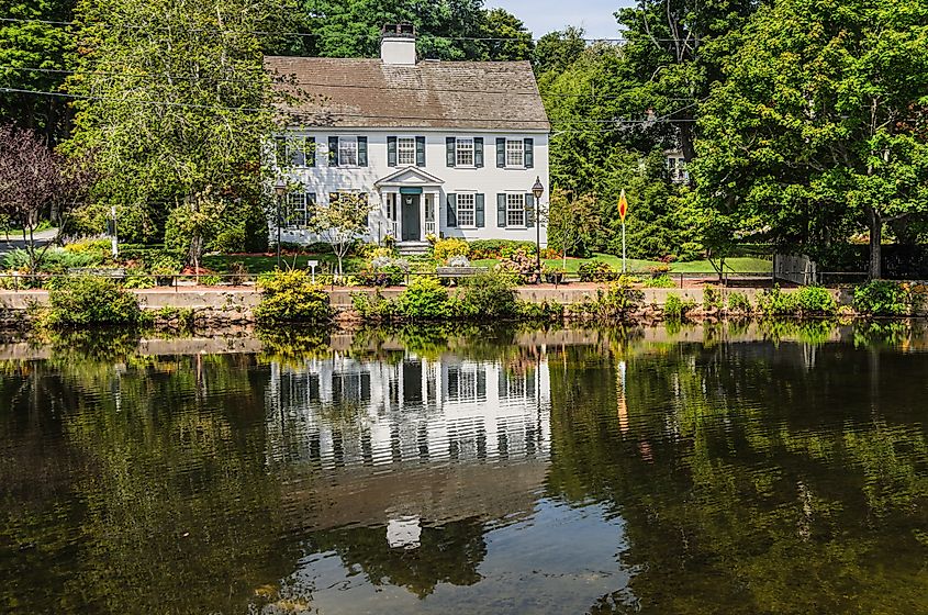 Classic white colonial house with green shutters beside a calm river, reflected in the water and surrounded by lush trees and garden landscaping.