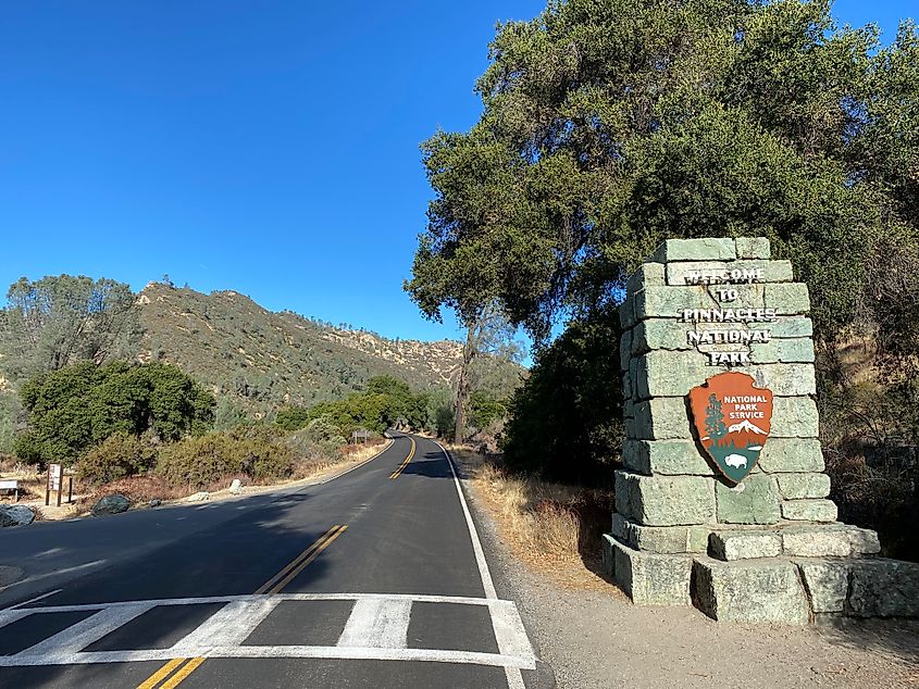 A rocky roadside pillar displays the Pinnacles National Park entrance sign. The paved road leads toward shrub-covered mountains.