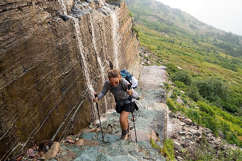 Brave woman hiker climbs up a narrow trail along a waterfall at the edge of a cliff, in Glacier National Park at Grinnell Glacier Trail.