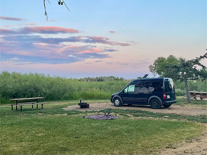 A small blue van sits at a prairie campground under pink/purple evening clouds.