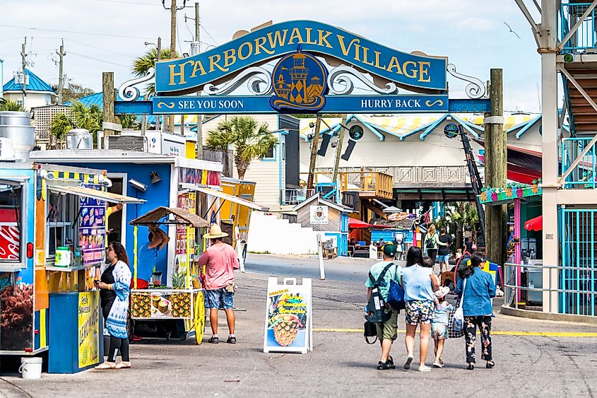 Harborwalk Village sign in Destin, Florida, USA, with people walking, shopping, and dining at cafes and street vendor restaurants.