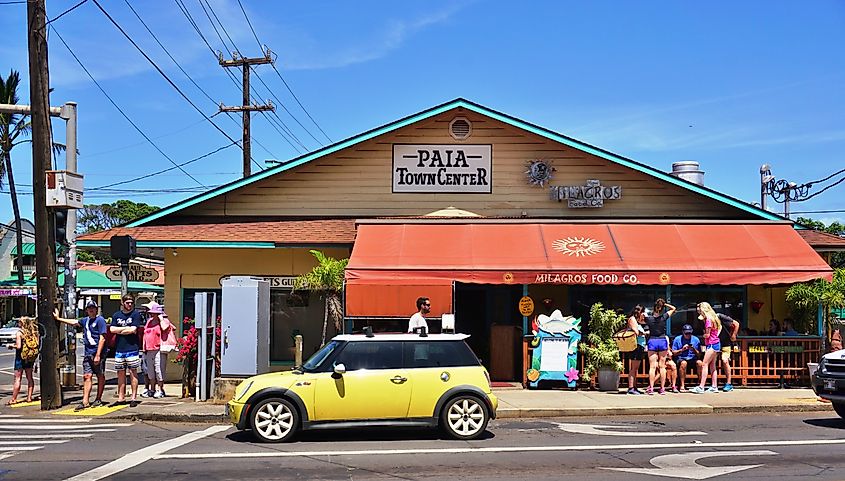 People gather outside Paia Town Center and Milagros Food Co. in Paia, Maui, with a yellow Mini Cooper parked along the street under a clear blue sky.