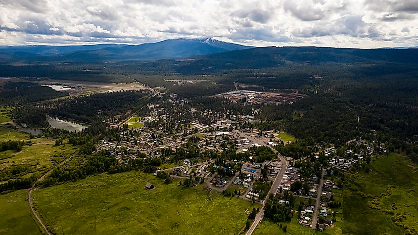 Aerial view of a small town surrounded by lush greenery, forests, and distant mountains under a cloudy sky. The scene conveys a peaceful, rural atmosphere.