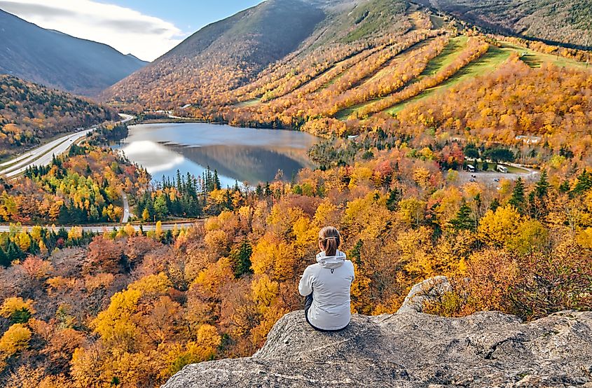 Woman hiking at Artist's Bluff in autumn, with a view of Echo Lake and vibrant fall colors in Franconia Notch State Park, White Mountain National Forest, New Hampshire