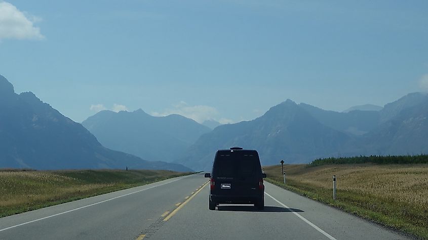 A small blue van drives on an empty mountainous highway. 